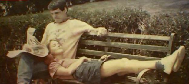 A man reading a newspaper on a park bench, with another person lying down and resting their head on his lap.
