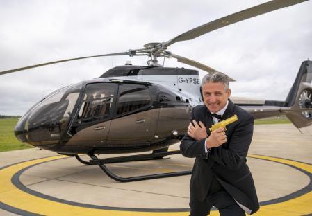 Man in tuxedo holding a gold gun in front of a helicopter.