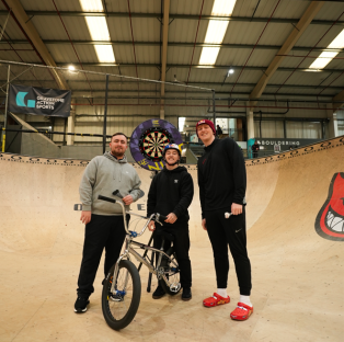 Three men posing with a BMX bike in a skate park.