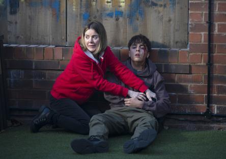 A woman cradles a bleeding teenage boy against a brick wall.